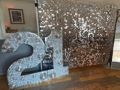 A silver glitter ball 21st birthday party decoration set featuring a large decorative number 2, a matching number 1, and a large square sequin backdrop, set on a wooden floor in an indoor venue.